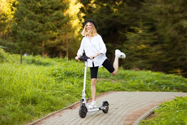 Chica hermosa alegre en sombrero negro y camisa blanca montando un scooter eléctrico en el parque de verano. Alquiler de vespa, libertad de movimiento —  Fotos de Stock