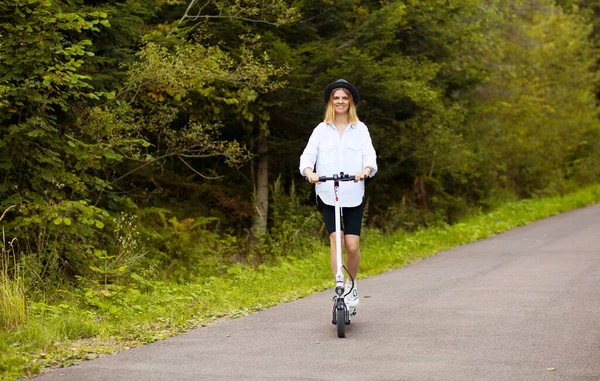 Vidám, gyönyörű lány fekete kalapban és fehér ingben, elektromos robogóval a nyári parkban. Scooter kölcsönzés, a mozgás szabadsága — Stock Fotó