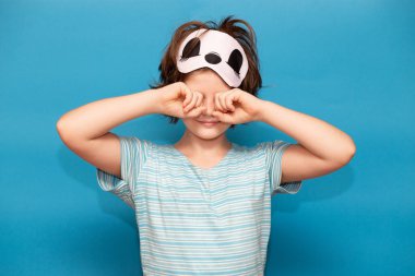 Uyku maskesi takmış yorgun bir kızın portresi. Mavi üzerine izole bir şekilde uyuduktan sonra ağlayıp gözlerini ovalıyor. Dişi çocuk uyanmak ve okula gitmek istemiyor. Sabah konsepti.