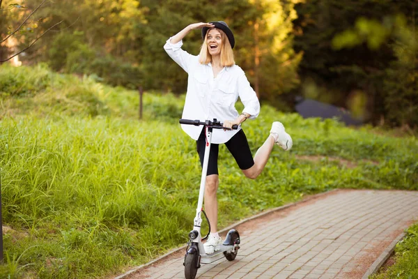 Vidám, gyönyörű lány fekete kalapban és fehér ingben, elektromos robogóval a nyári parkban. Scooter kölcsönzés, a mozgás szabadsága — Stock Fotó