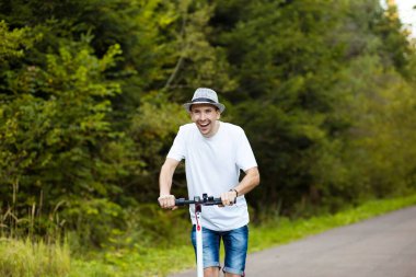 Elektrikli taşıma. Beyaz tişörtlü, güneş gözlüklü ve şapkalı genç yakışıklı adam güneşli bir yaz gününde elektrikli scooter bisikletini sürmeye hazır. Ekolojik ulaşım kavramı.