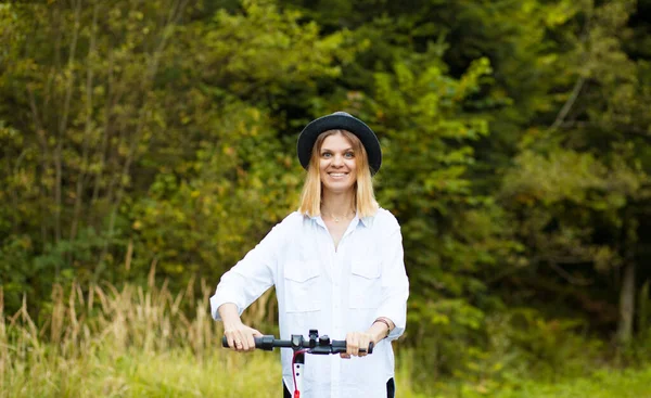 Trendy unbeschwerte Frau in weißem Hemd und schwarzem Hut fahren auf Tretroller Outdoor in der sommerlichen Parkstraße. Ökologisches Elektroverkehrskonzept. — Stockfoto