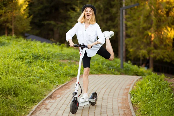 Vidám, gyönyörű lány fekete kalapban és fehér ingben, elektromos robogóval a nyári parkban. Scooter kölcsönzés, a mozgás szabadsága — Stock Fotó