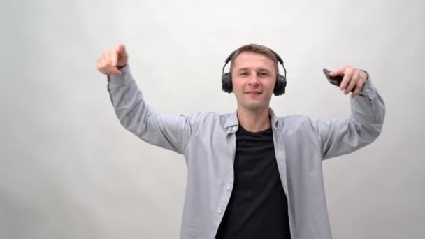 Mladý muž na bílém pozadí ve sluchátkách a smartphonu poslouchá hudbu a tance. Veselý emocemi, evropského původu. — Stock video