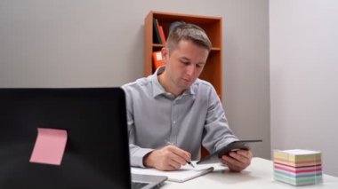 Genç bir ofis çalışanı elinde bir tablet tutuyor, uzaktan kumandayla. Bilgisayarın başında oturuyor..