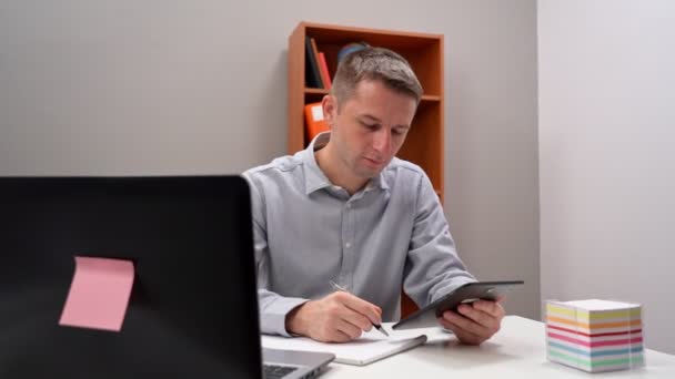 Νεαρός υπάλληλος γραφείου που κρατάει ένα τάμπλετ στα χέρια του, σε απομακρυσμένη εργασία. Καθισμένος στον υπολογιστή. — Αρχείο Βίντεο
