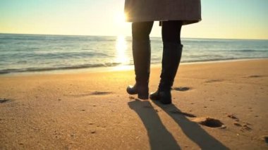 Kadın figürü, belden aşağısı, sahilde uzun botlar giyerek yürüyor. Yavaş çekim..