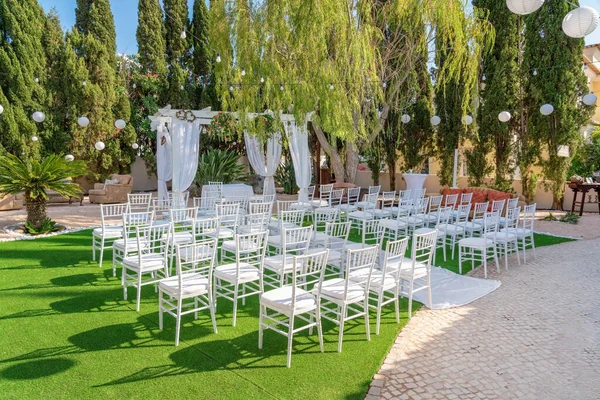 Venku svatební obřad nastavení s bílými židlemi v zahradě se stromy v pozadí. — Stock fotografie