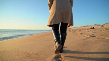 Kadın figürü, belden aşağısı, sahilde uzun botlar giyerek yürüyor. Yavaş çekim..