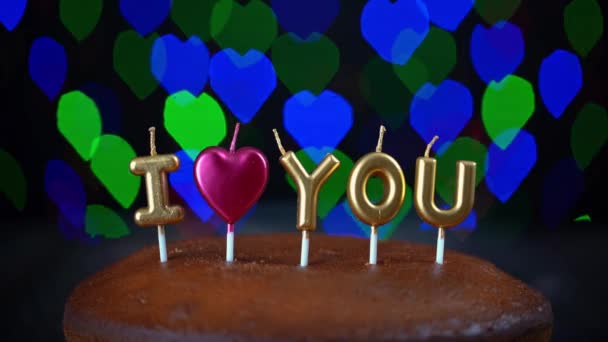 Video shot of simple Valentines Day cake with lit i love you candles, with a colorful background. In the background, blurry lights, bokeh of hearts blink. — Stock Video