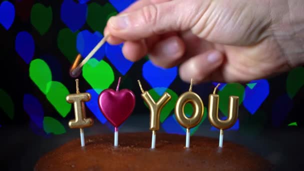 Mano, enciende velas-letras, te amo, en el pastel, con un fondo en un bokeh parpadeante en forma de corazón. El día de San Valentín. — Vídeos de Stock