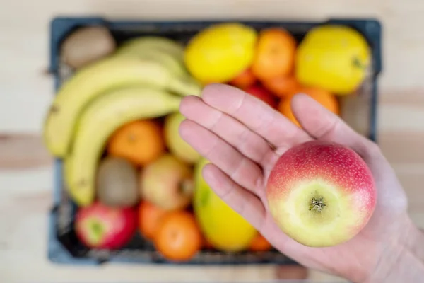 Hand holding an apple in focus and beneath it, is a basket full of varied fruit out of focus. — Stock Photo, Image