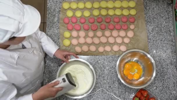 Baker morrande äggvita med en elvisp. Några okokt makaroner, en skål med gulor i en skål och jordgubbar kan också ses på bordet. — Stockvideo