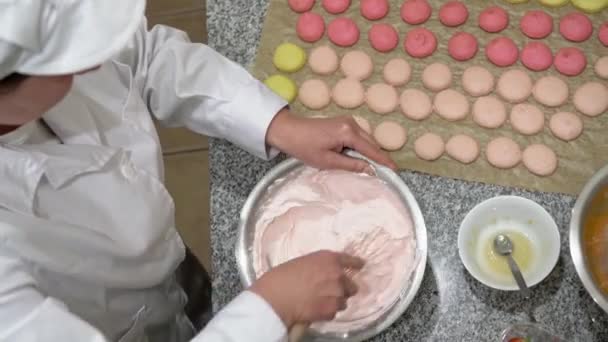 Bovenaanzicht van een bakker, met de hand kloppend wat slagroom in een metalen kom. Veel macarons zijn ook te zien op de tafel. — Stockvideo