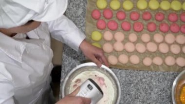 Fırıncı elektrikli çırpıcıyla yumurta beyazı çırpıyor. Masada pişmemiş makaronlar, bir kase yumurta sarısı ve çilek de görülebilir..