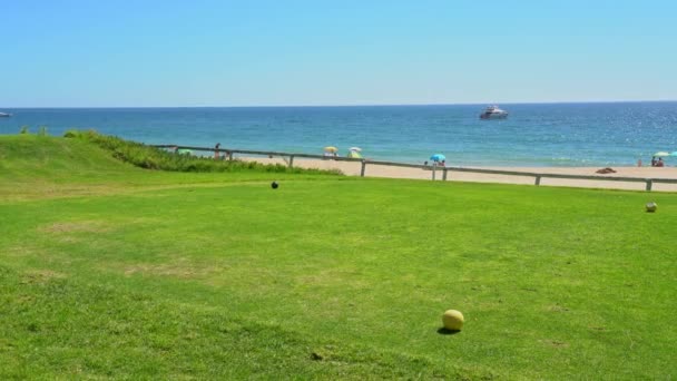 Όραση ενός χλοερό γήπεδο γκολφ, ένα καθαρό γαλάζιο ωκεανό με ένα γιοτ στο βάθος και μερικοί άνθρωποι στην παραλία. Vale do Lobo, Πορτογαλία. — Αρχείο Βίντεο