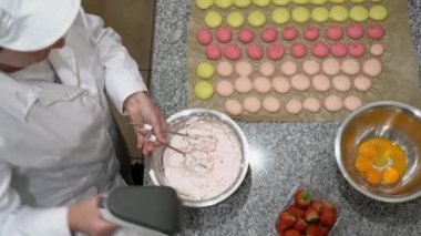 Fırıncı elektrikli çırpıcıyla yumurta beyazı çırpıyor. Masada pişmemiş makaronlar, bir kase yumurta sarısı ve çilek de görülebilir..