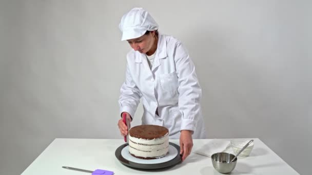 Baker verwijdert de overtollige room van de zijkanten van een chocoladetaart door de standaard te draaien en een spatel te gebruiken. — Stockvideo