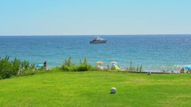 Çimenli bir golf sahası, açık mavi bir okyanus, uzakta bir yat ve sahilde bazı insanlar. Vale do Lobo, Portekiz.