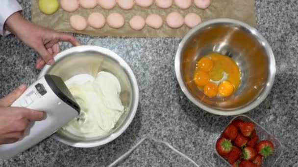 Baker klopte eiwit met een elektrische klopper. Sommige ongekookte macarons, een kom dooiers in een kom en aardbeien zijn ook te zien op de tafel. — Stockvideo