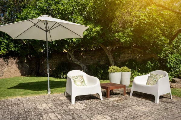 Зелений сад з парою білих стільців, маленький стіл і велика парасолька, щоб забезпечити тінь в яскравий сонячний день . — стокове фото