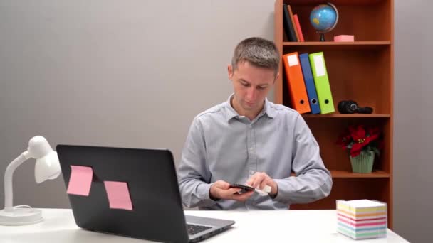 Jovem, em seu trabalho no escritório, limpando seu dispositivo de smartphone. Há também um computador portátil colocado em sua mesa. — Vídeo de Stock