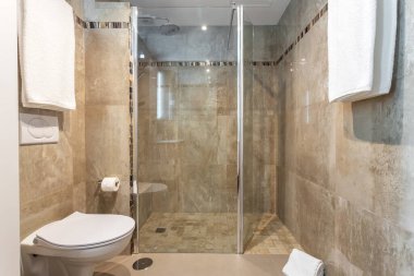 Bej renkli banyo iç kısmı, cam kapılı bir duş ve seramik tuvalet, ayrıca duvarlarda birkaç havlu..