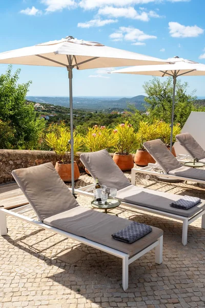 Liegestühle mit Sonnenschirmen als Schattenspender, eine Flasche Wein auf einem kleinen Tisch und eine schöne Aussicht auf die Berge an einem klaren, sonnigen Tag. Stockfoto