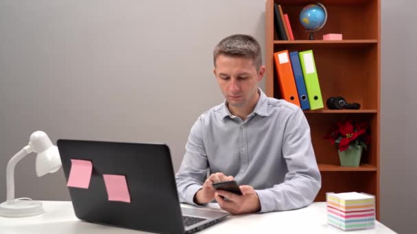 Entreprenör multitasking i en kontorsmiljö genom att använda en smartphone och en bärbar dator för att forska. Sitter vid ett skrivbord med en bokhylla bakom sig. — Stockvideo