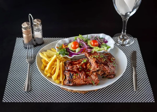 Glazed barbecue ribs with fries and a healthy salad. Cutlery and a glass of water, set up for a meal.