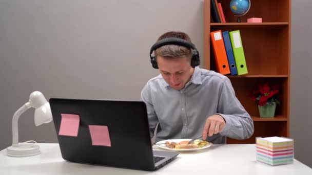 Ung kontorsarbetare, student, föreståndare, med hörlurar, äter sin lunch på hemmakontoret, under - en pandemi. Med en dator i förgrunden. — Stockvideo
