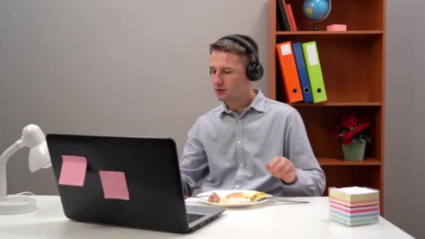 Jovem funcionário de escritório, estudante, gerente, usando fones de ouvido, comendo seu almoço em seu escritório em casa, durante uma pandemia. Com um computador em primeiro plano. — Vídeo de Stock