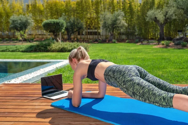 Donna sta praticando ginnastica fitness, pilates, all'aperto vicino casa e piscina, lezioni online con un allenatore. Con uno sfondo al giardino. — Foto Stock