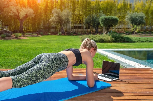 Donna sta praticando ginnastica fitness, pilates, all'aperto vicino casa e piscina, lezioni online con un allenatore. Con uno sfondo al giardino. — Foto Stock