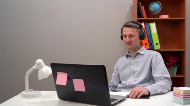 Un joven trabajador se comunica con un cliente en una conferencia en línea desde la oficina, a través de auriculares. — Vídeos de Stock