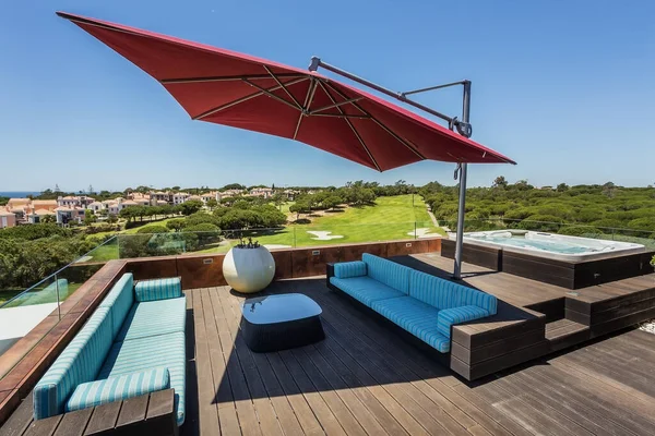 Moderna terraza con un par de sofás un paraguas y un jacuzzi en el lado, detrás de una increíble vista de la zona verde cubierta de hierba. — Foto de Stock