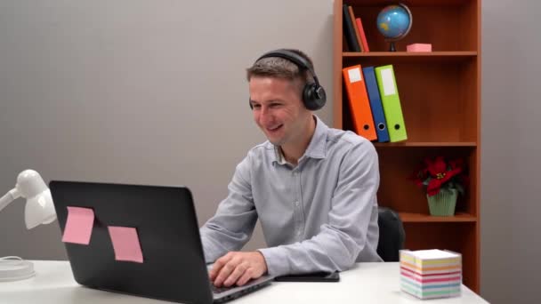 Un joven trabajador se comunica con un cliente en una conferencia en línea desde la oficina, a través de auriculares. — Vídeos de Stock