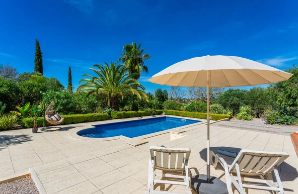 Pool mit Sonnenschirm und Liegestühlen am Pool. Klarer blauer Himmel und schöne Bäume im Hintergrund an einem schönen Sommertag. — Stockfoto