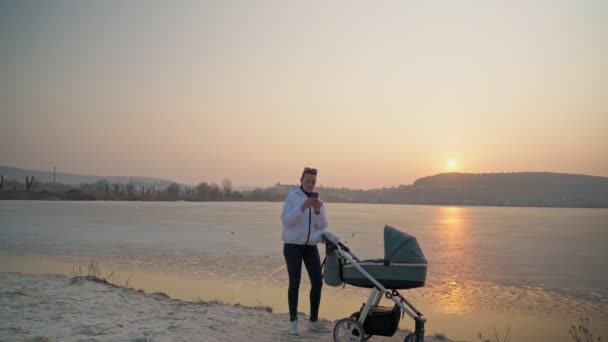 Eine junge Frau mit Kinderwagen und neugeborenem Baby geht auf einem Quellsee spazieren. Bei Sonnenuntergang. Mütterliche Gefühle und Gefühle. — Stockvideo