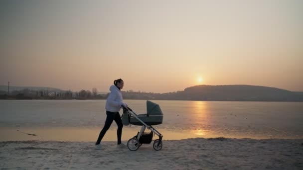 En ung kvinde med klapvogn og en nyfødt baby, der går på en forårssø. Under solnedgang. Moderens følelser og følelser. – Stock-video