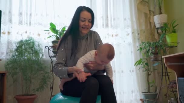 Uma jovem mãe na sala de estar, fazendo ginástica com o bebê, reabilitação, em uma bola de ginástica. — Vídeo de Stock