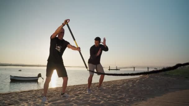 Homme faisant un entraînement de mouvement d'aviron sur une plage de sable, avec une bande élastique accompagnée de son entraîneur personnel. Imitation d'aviron sur une planche de surf. Faro Portugal 2021 16 avril — Video