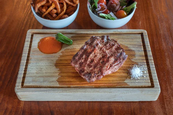 Um pedaço grelhado de bife fresco, médio raro. Em um conselho de textura e verduras no escritório traseiro. Molho de pimenta e sal, para o melhor sabor. — Fotografia de Stock