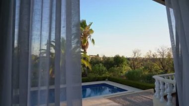 Modern oturma odasının perdeleri rüzgarda uçuşan görüntüsü bir balkon ve dış havuzu ortaya çıkarıyor. Ve tropik bir bahçe.