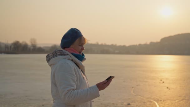 Una mujer mayor, bonita, de edad de jubilación, en la orilla del lago, se comunica a través de un teléfono inteligente por mensajes de voz. — Vídeo de stock