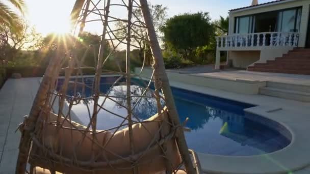 Lyxig hängande gungstol, bredvid den rena poolen och villaresidenset. Med vacker trädgård och himmel i sommarväder. Flytta kameran framåt. — Stockvideo