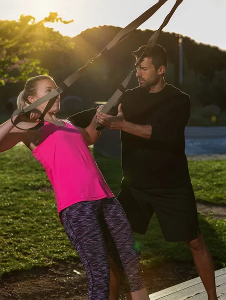 Osobní fitness trenér a mladá žena klient, opravuje cvičení s popruhy. Ráno v parku. Slunečný den. — Stock fotografie