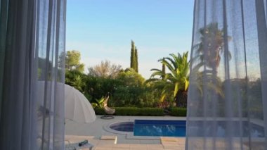 Modern oturma odasının perdeleri rüzgarda uçuşan görüntüsü bir balkon ve dış havuzu ortaya çıkarıyor. Ve tropik bir bahçe.