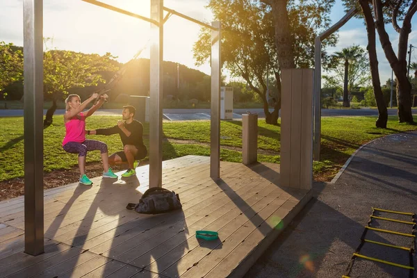 Kişisel fitness eğitmeni ve genç bayan müşteri, egzersizleri kayışlarla düzeltiyor. Sabah park yerinde. Güneşli bir gün. — Stok fotoğraf