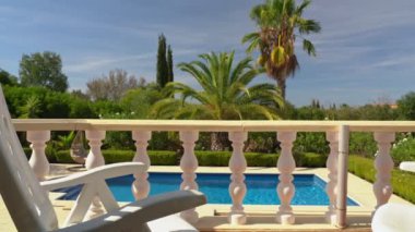 Kameranın soldan sağa, yandan sağa hareket etmesi, terasın taş sütunları, villanın balkonu, güzel havuza bakan. Turistler için palmiye ağaçları olan bir bahçe.